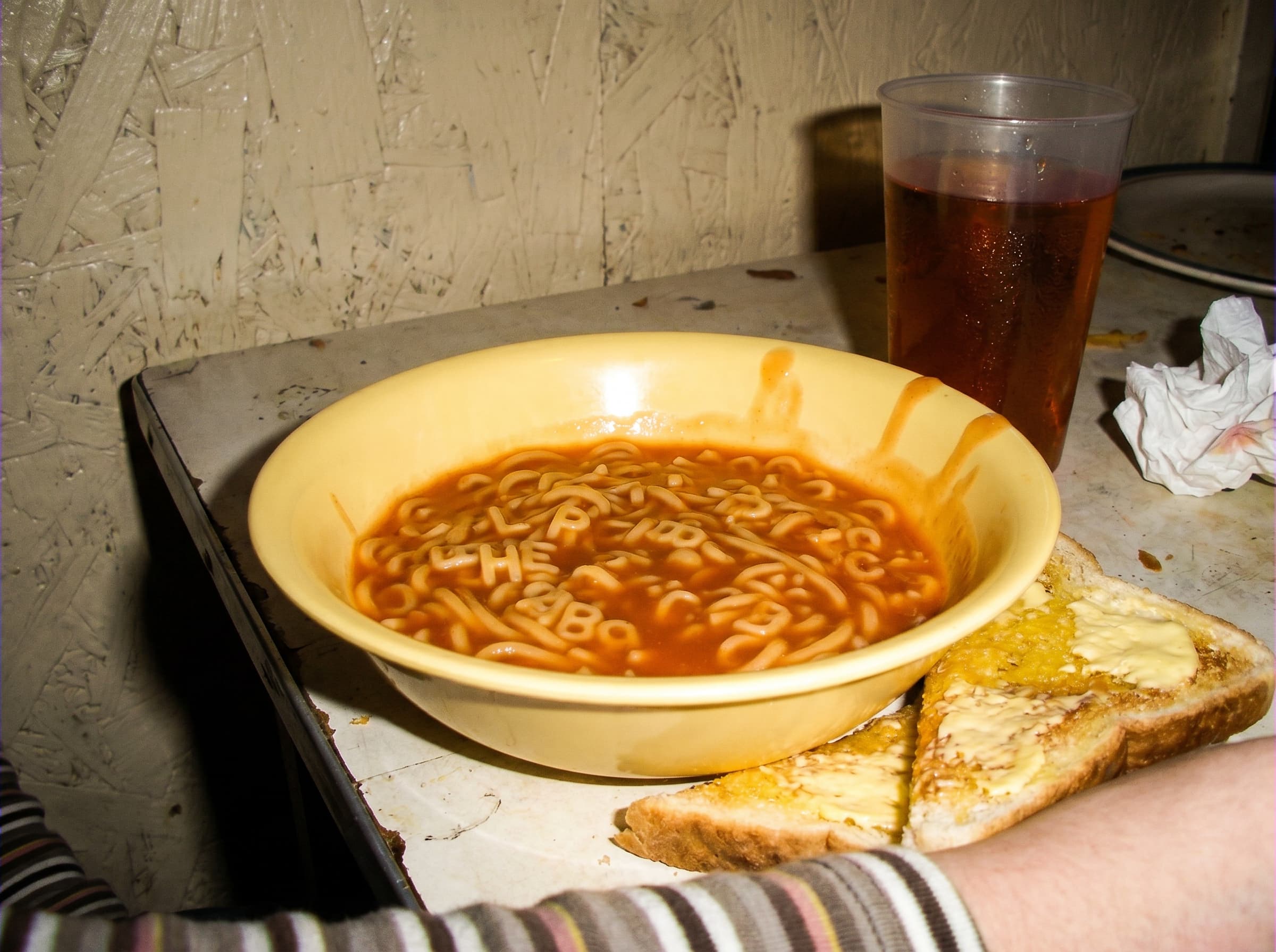 A bowl of tinned alphabet spaghetti spilling letters across a Formica diner table, lit by direct flash.